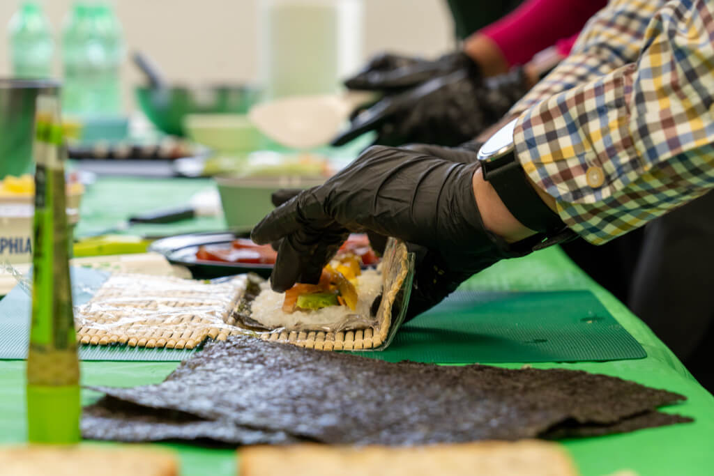 Primer plano de manos con guantes negros enrollando un maki de salmón y aguacate en actividad de team building